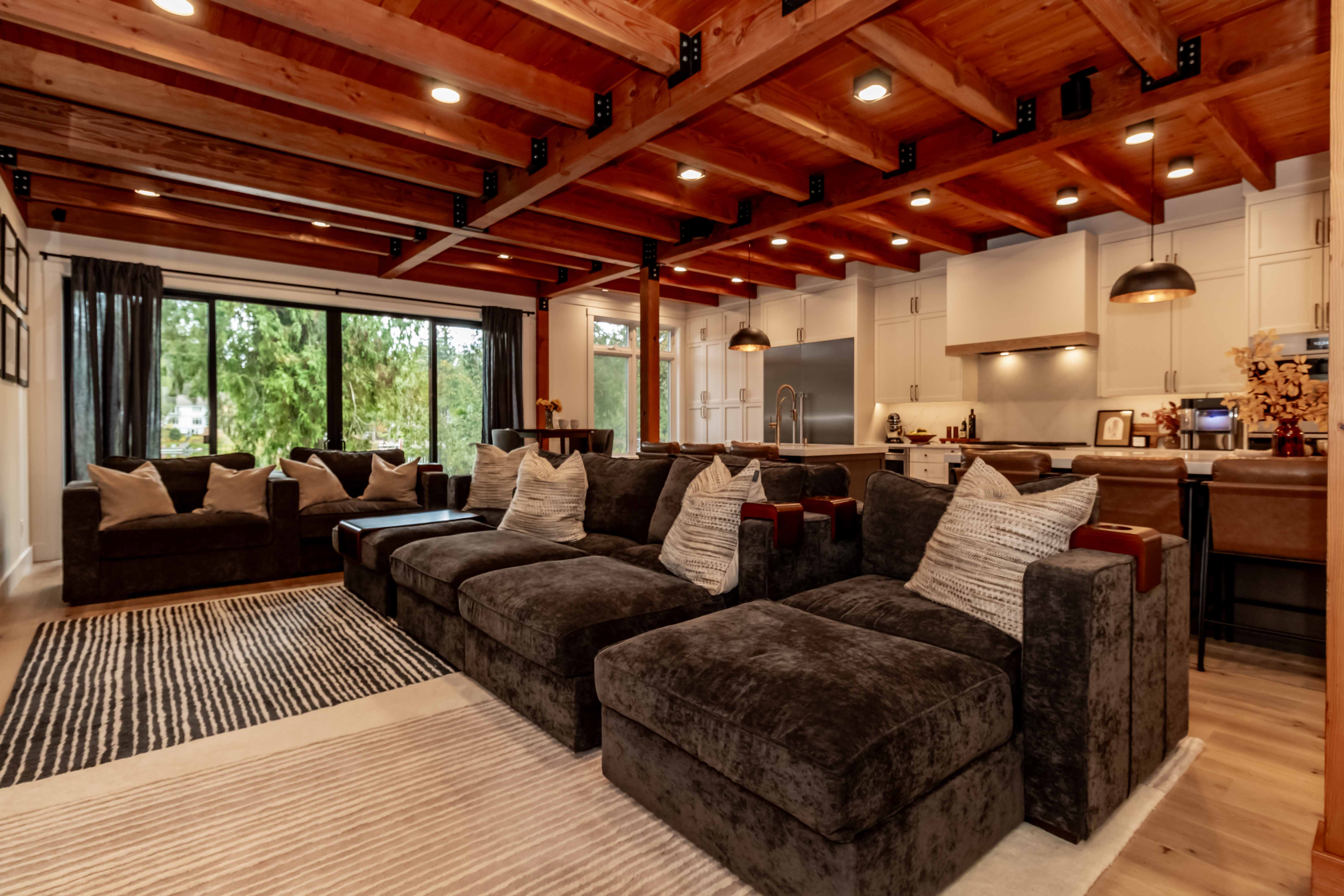 A modern open-concept living room and kitchen with exposed wooden ceiling beams, dark sectional sofas, large windows, and pendant lighting over a kitchen island.