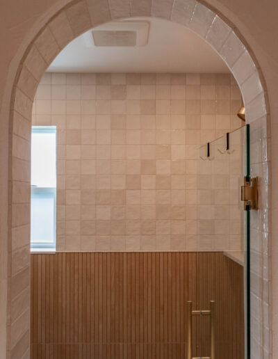 Arched doorway into a shower with square cream tile, a fluted wood-look lower wall and brass fixtures